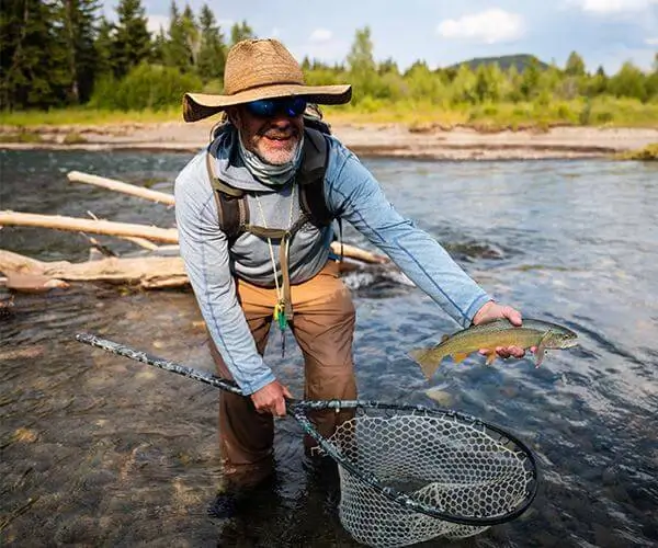 Man in a wide-brimmed hat holding a caught fish above a net, standing in a river surrounded by trees, representing guided fly fishing experiences in Jackson Hole, Wyoming.