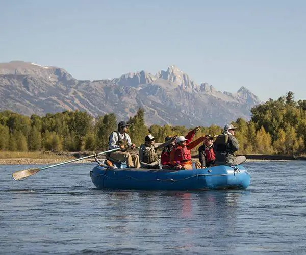 Group of anglers in a blue raft on a river, surrounded by the scenic mountains of Jackson Hole, Wyoming, highlighting guided fly fishing experiences.
